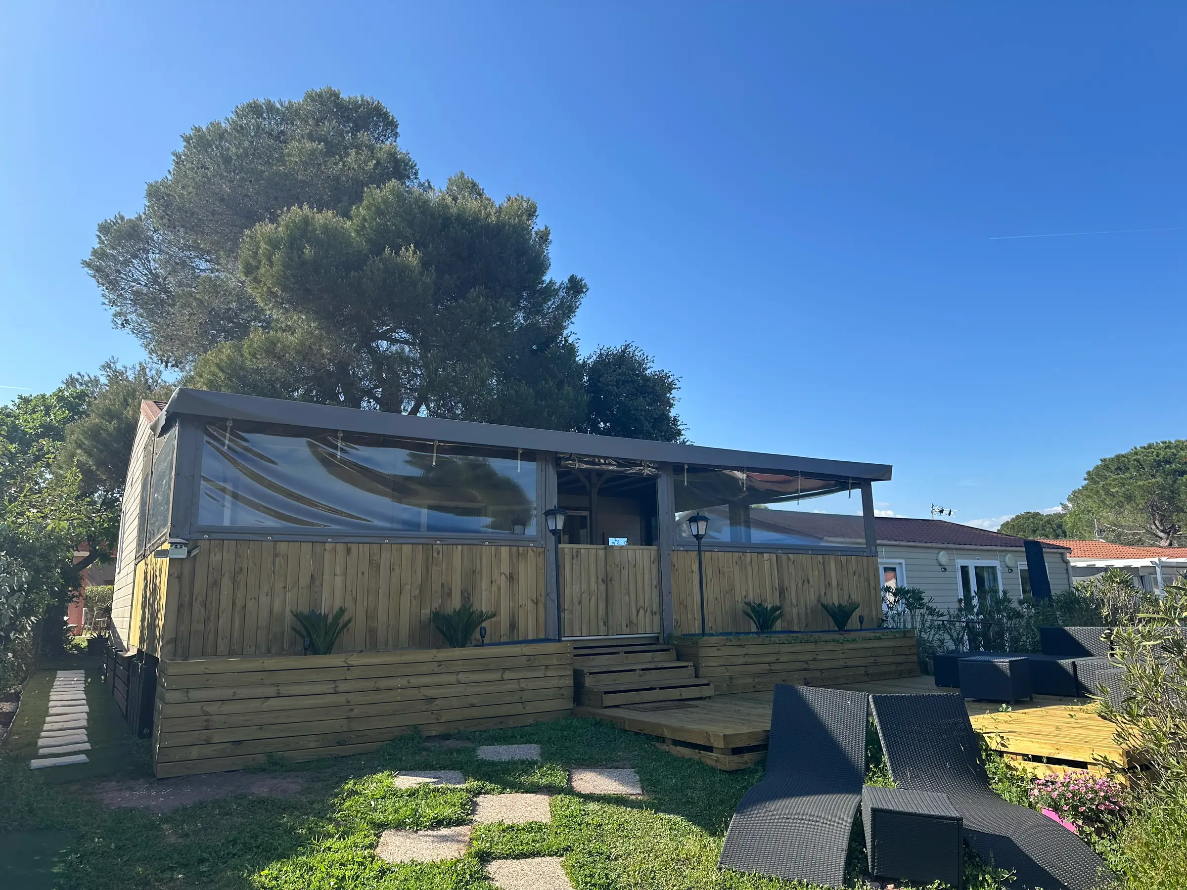 Façade du chalet et sa terrasse close en bois clair sous un pin parasol